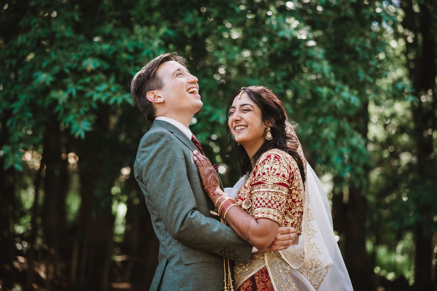 A couple shares a joyful moment in a forest setting, with one wearing traditional South Asian wedding attire.