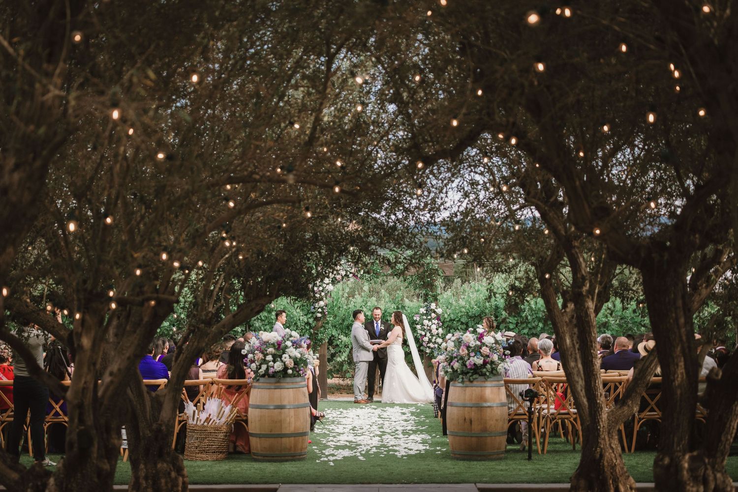 Outdoor wedding ceremony setup with wine barrels and twinkle lights creating romantic atmosphere.