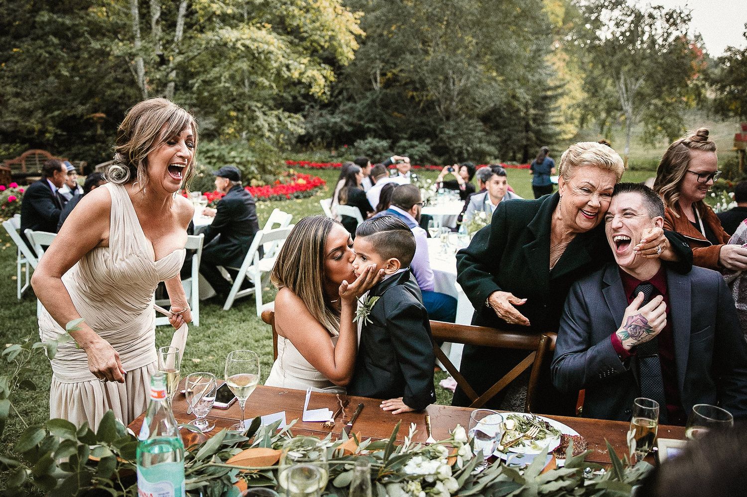 Wedding guests share joyful laughter during an outdoor reception celebration in a garden setting.