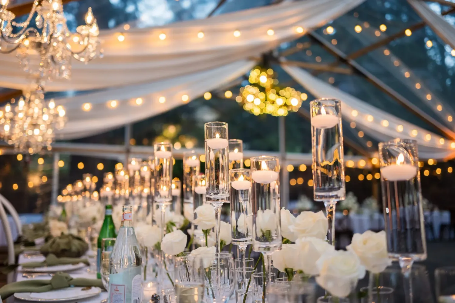 Elegant wedding reception tent decorated with crystal chandeliers, string lights, and white floral centerpieces.