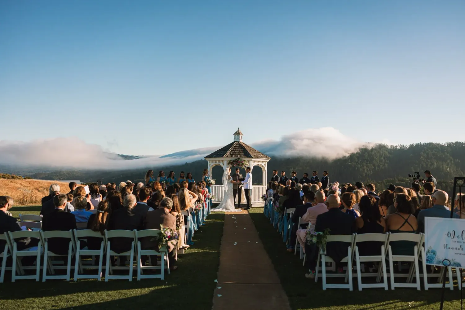 Panoramic view of an outdoor wedding ceremony setup with white chairs and mountain landscape backdrop at sunset.