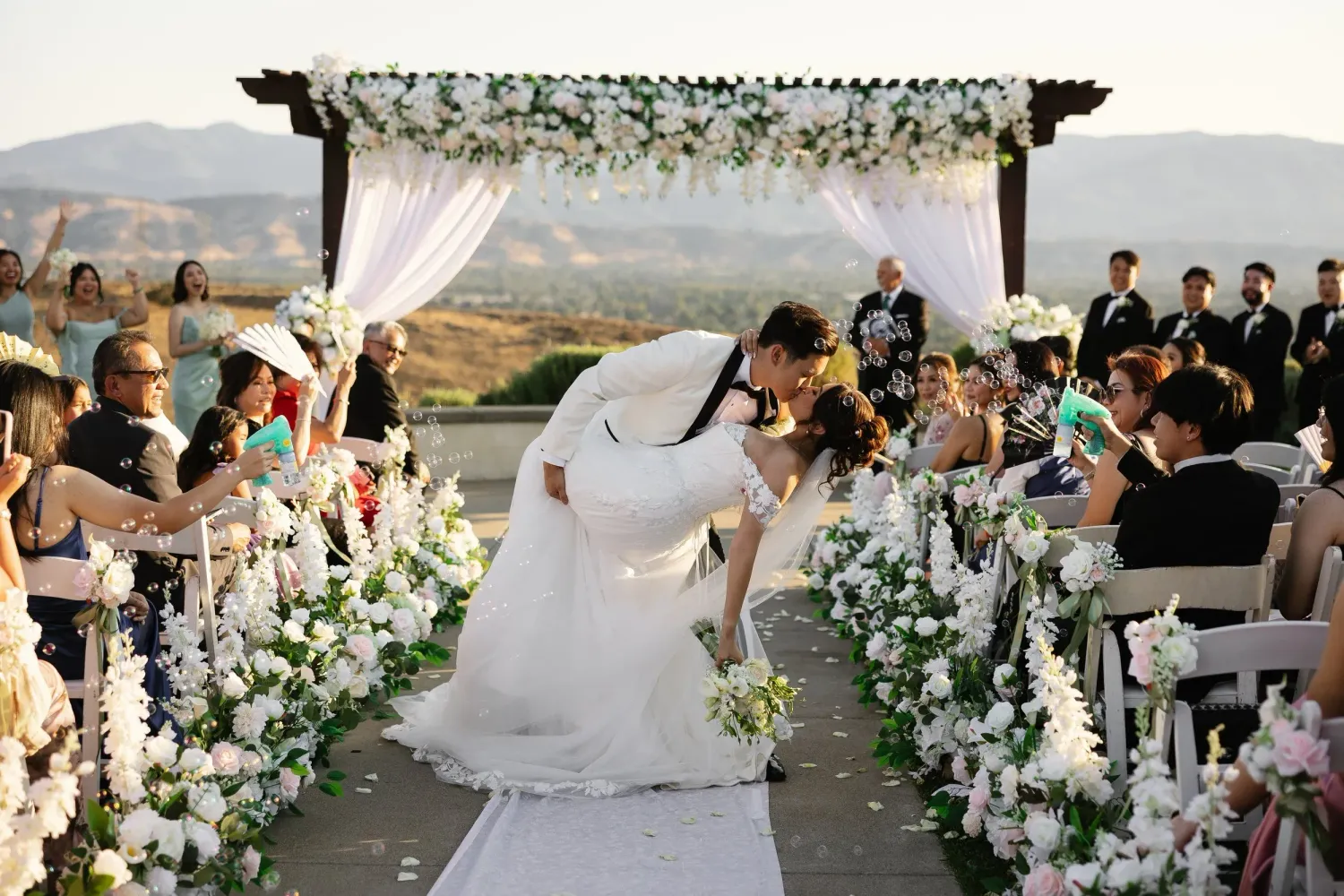 A wedding ceremony setup with white floral arrangements and mountain views in an outdoor venue.