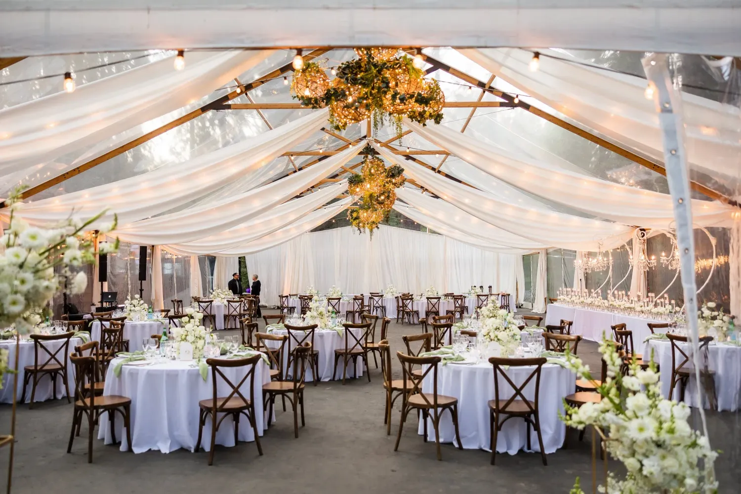 Elegant wedding reception setup under white draped tent with chandeliers and round tables with cross-back chairs.