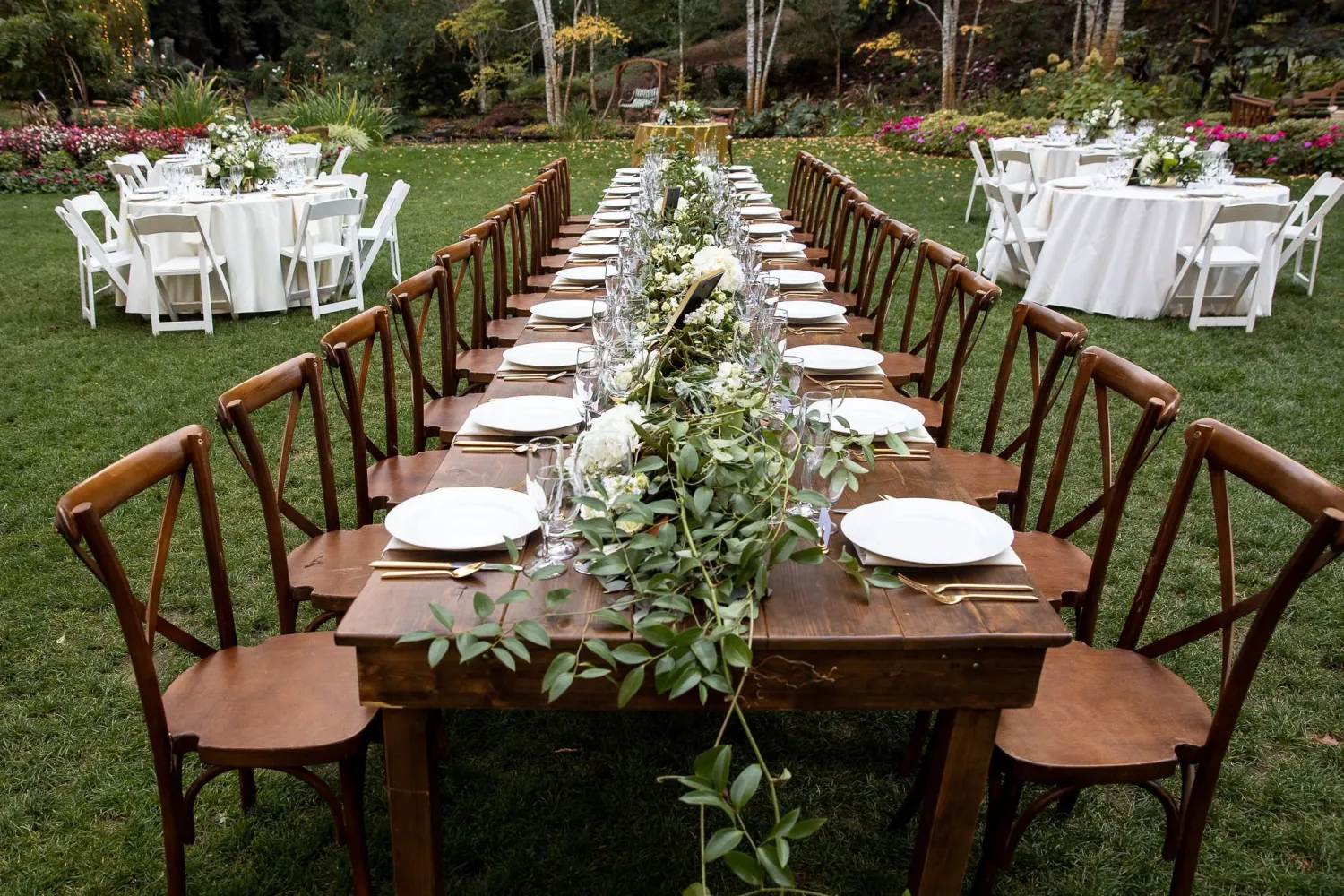 Elegant outdoor wedding reception setup with wooden cross-back chairs and white tablecloths under trees.