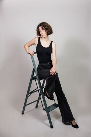 A model wearing black posing on a green step ladder against a grey background. Model