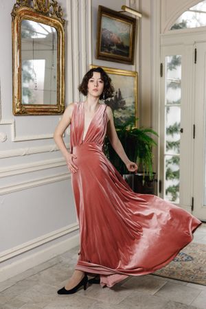 A model in a flowing pink velvet dress poses in an elegant white room with gold-framed artwork. Model