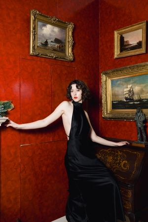 A dramatic portrait of a model in black dress against red walls with vintage sailing ship paintings. Model