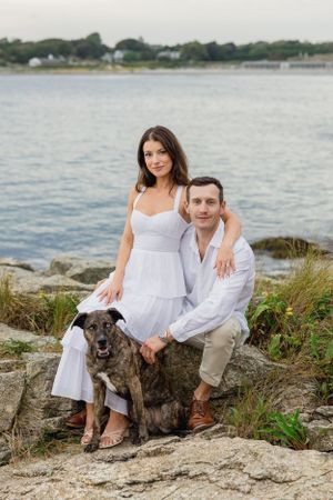 A couple and their brindle dog pose together on rocky coastal terrain with water views. Cliff Walk, Newport, RI