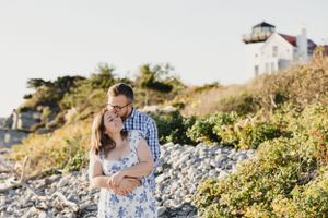 A couple embraces on a rocky coastal path with a lighthouse visible in the background. Engagement Session RI