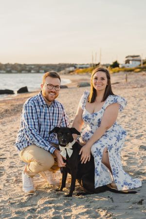 A couple poses with their black dog on a sandy beach during sunset. Engagement Session RI
