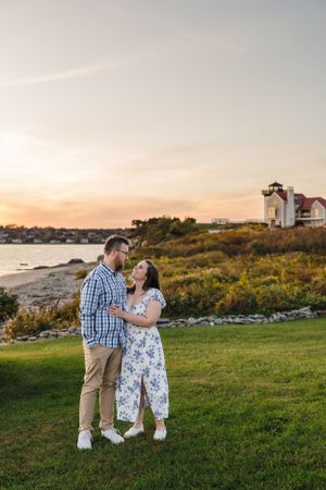 A couple shares a romantic moment on a grassy hill with a lighthouse and sunset in the background. Engagement Session RI