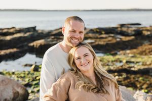 A couple smiles together by rocky coastal waters during sunset. Engagement Session Newport, RI