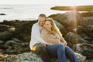 A couple sits together on coastal rocks during golden hour. Engagement Session Newport, RI