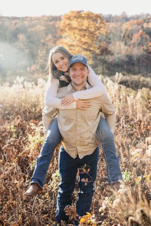 A couple shares a playful piggyback ride moment in a golden autumn field at sunset. Engagement Session CT