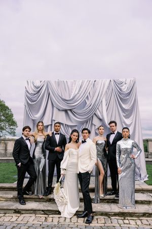 Wedding party in formal attire poses against silver draped backdrop outdoors.