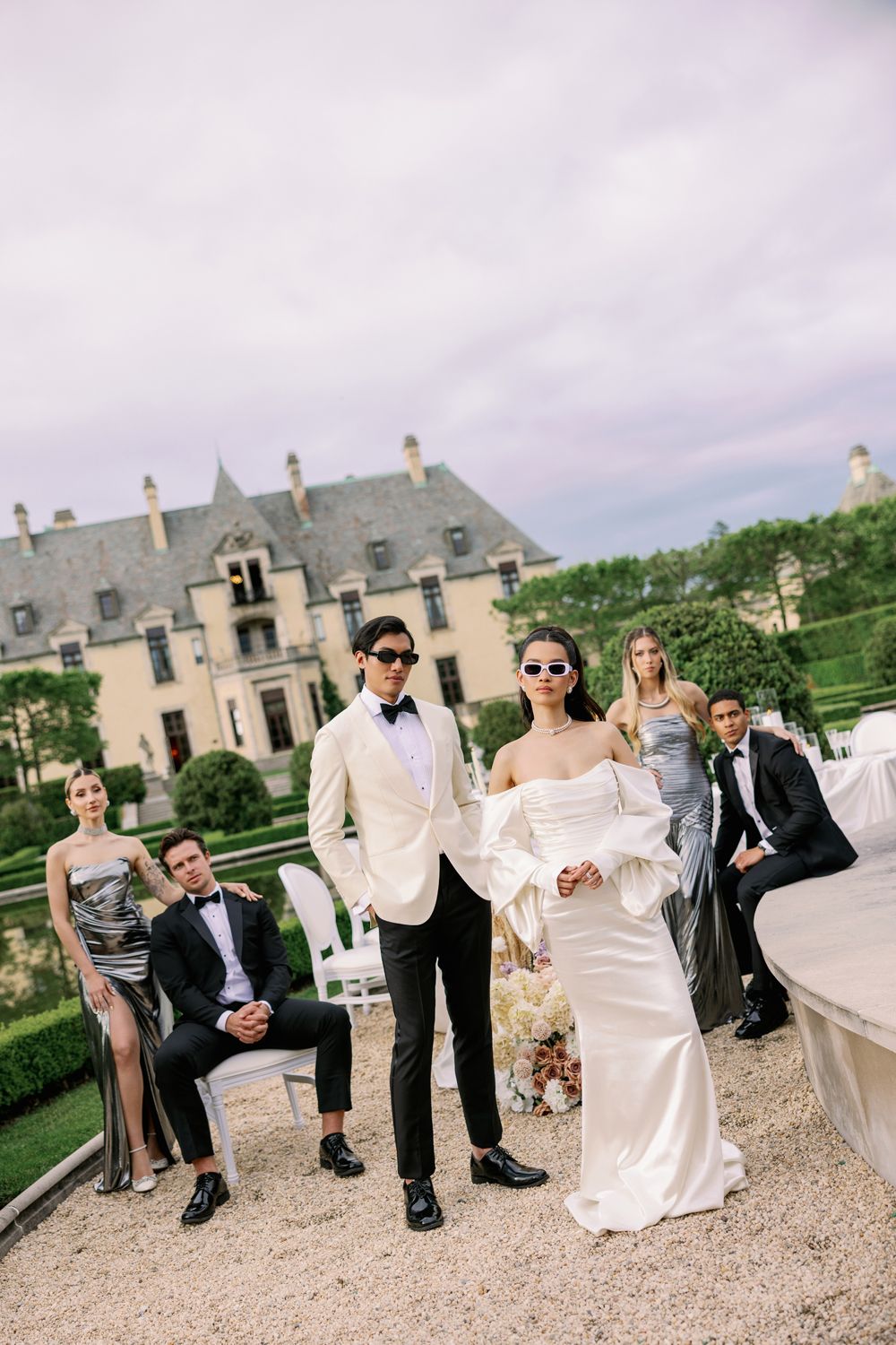 Wedding party members relax and socialize on stone steps outside an elegant chateau during an outdoor celebration.