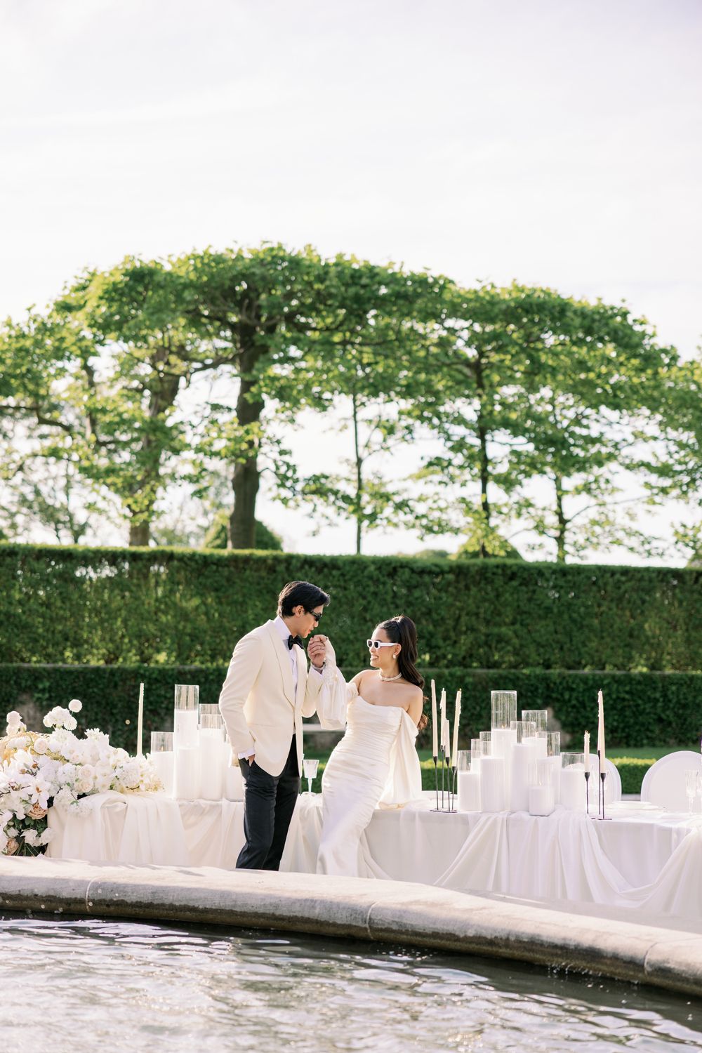 Elegant wedding reception setup with white tablecloths and decor along a waterfront terrace with manicured hedges.