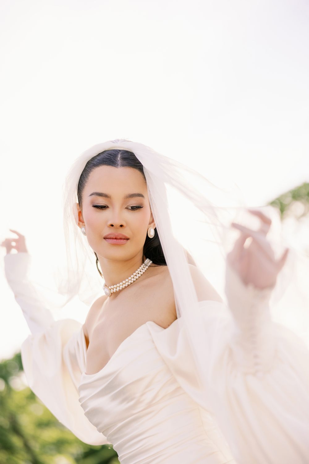 A bride in a flowing white dress and pearl necklace twirls gracefully against a soft outdoor background.