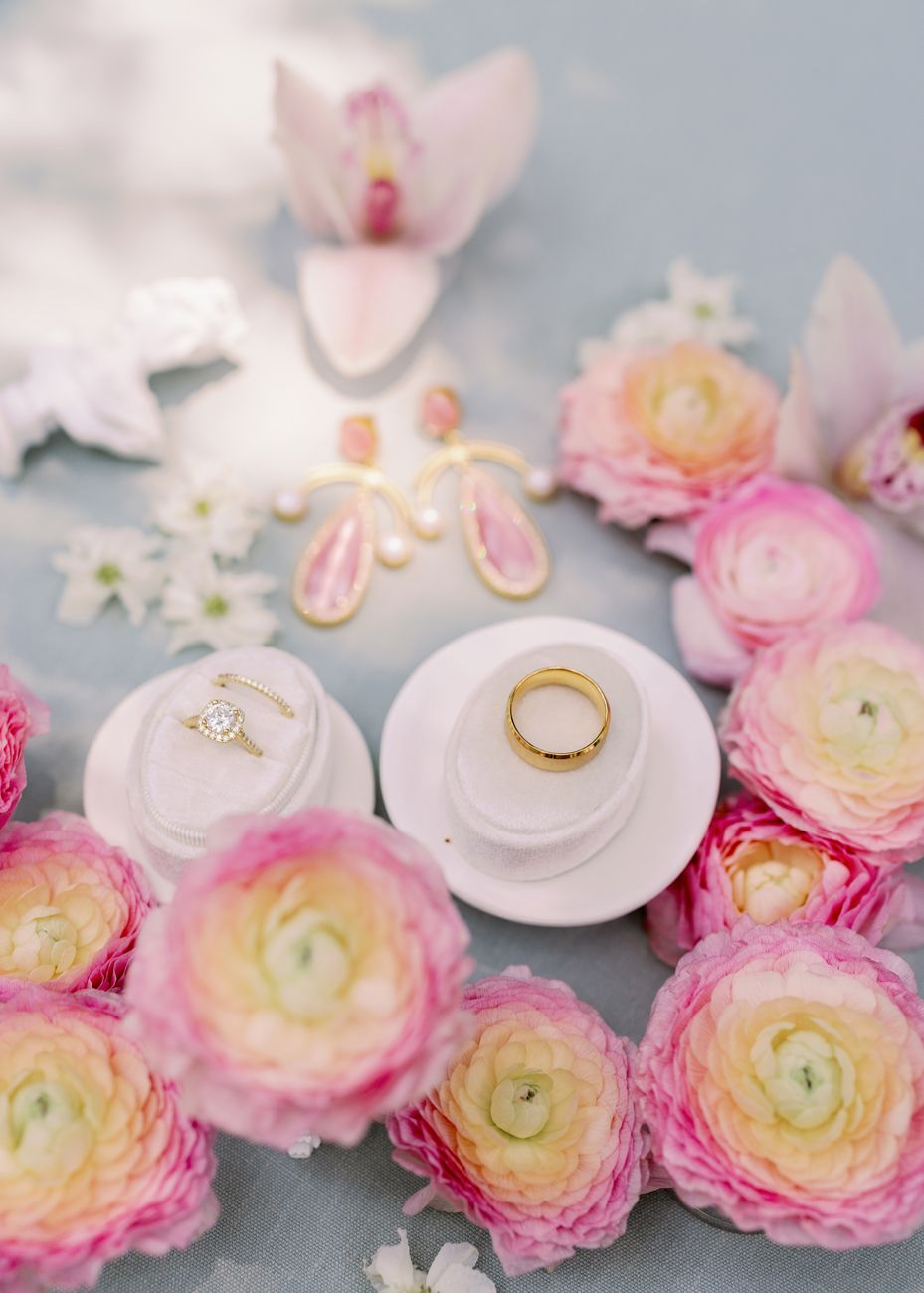 Wedding rings on white ceramic dishes surrounded by pink and peach ranunculus flowers.
