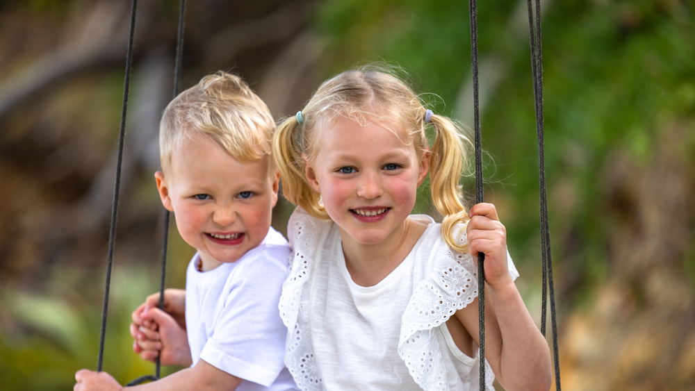 Two beautiful young siblings, one girl one boy sitting together on a swing at the beach with a green foliage background