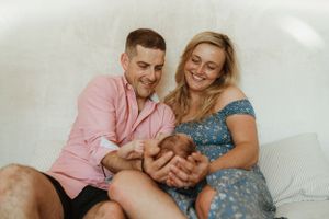 A couple in pink and blue outfits sharing a tender moment with their newborn on a couch.