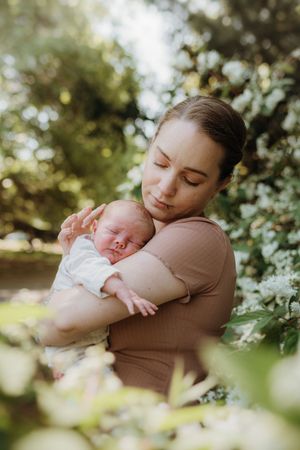 Mother cradling sleeping newborn baby among blooming white flowers outdoors.