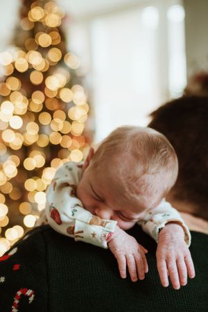 A sleeping infant in holiday clothing rests against dark fabric with Christmas lights in the background.