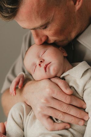 A tender moment captured as a parent gently kisses their sleeping newborn baby wrapped in a cream-colored sweater.
