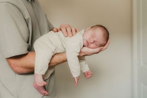 A sleeping newborn baby in white onesie rests peacefully on adult's forearms against neutral background.