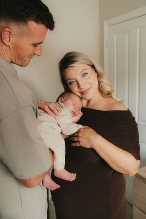 A loving couple holds their newborn baby in a cozy indoor setting with warm lighting and neutral tones.