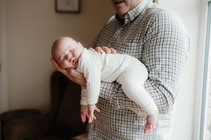Parent in plaid shirt holds sleeping baby wrapped in white blanket while standing in sunlit room.