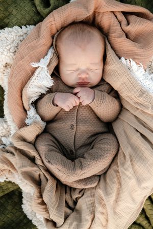 A sleeping newborn baby wearing a brown knit outfit lies swaddled in soft beige muslin fabric and white lace.