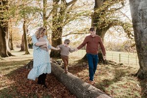 A family enjoys a playful moment balancing together on a fallen tree trunk in an autumn forest setting.