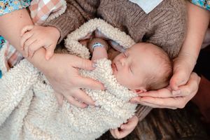 A sleeping newborn wrapped in a cozy cream knit blanket is gently cradled by caring hands.