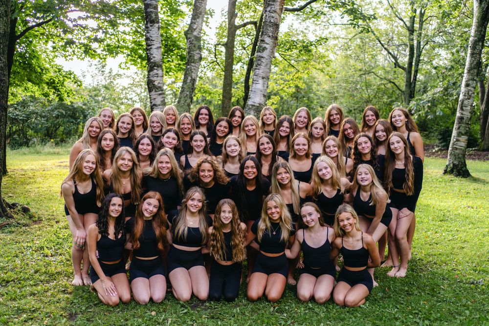 Large group wearing black outfits poses together outdoors in a wooded setting with birch trees and green grass.