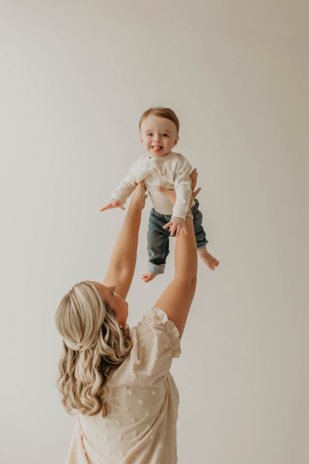 A joyful child wearing a beige sweater and jeans is lifted playfully in the air against a minimalist white background.