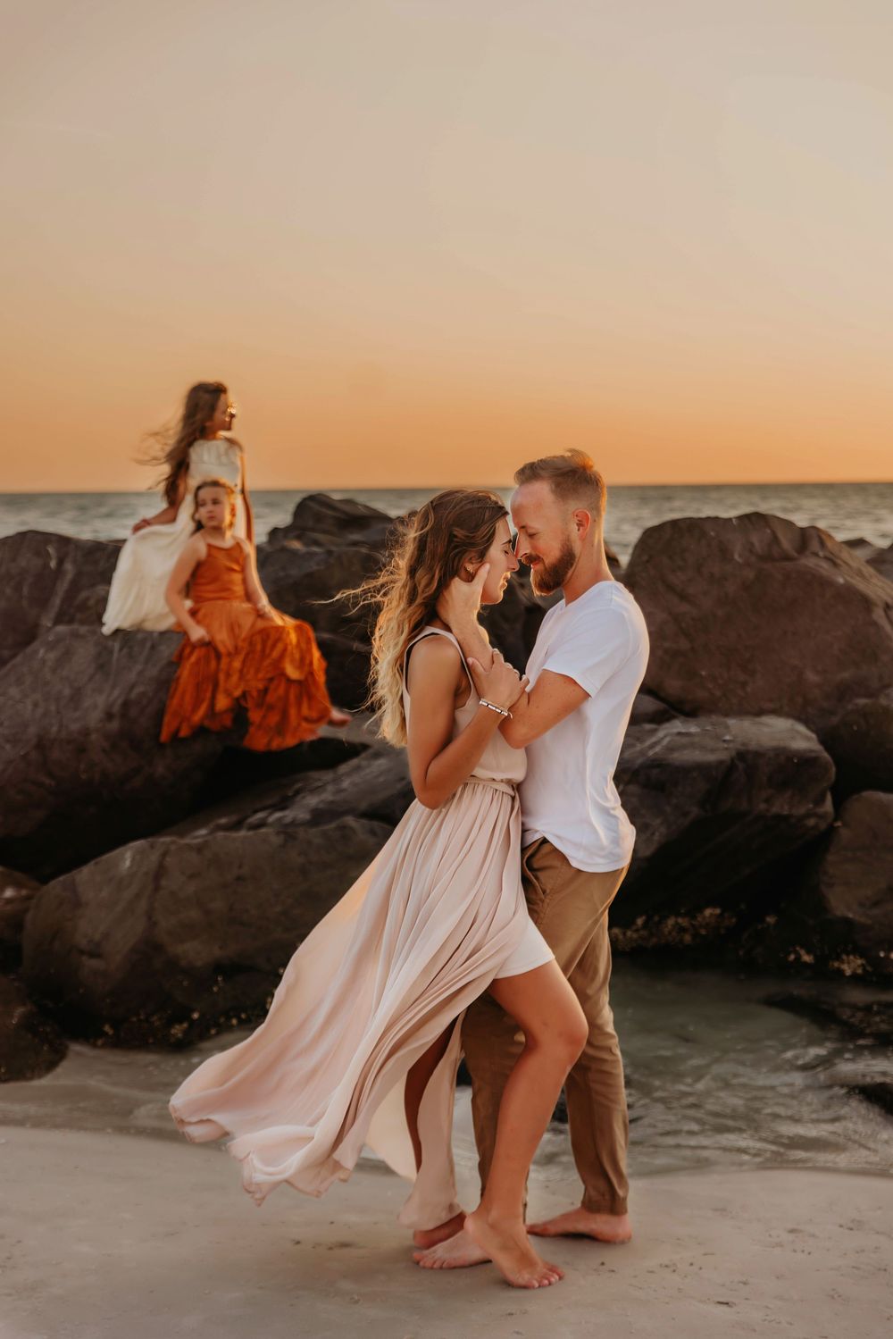 Three people dance along a rocky beach at sunset with flowing dresses and romantic lighting.