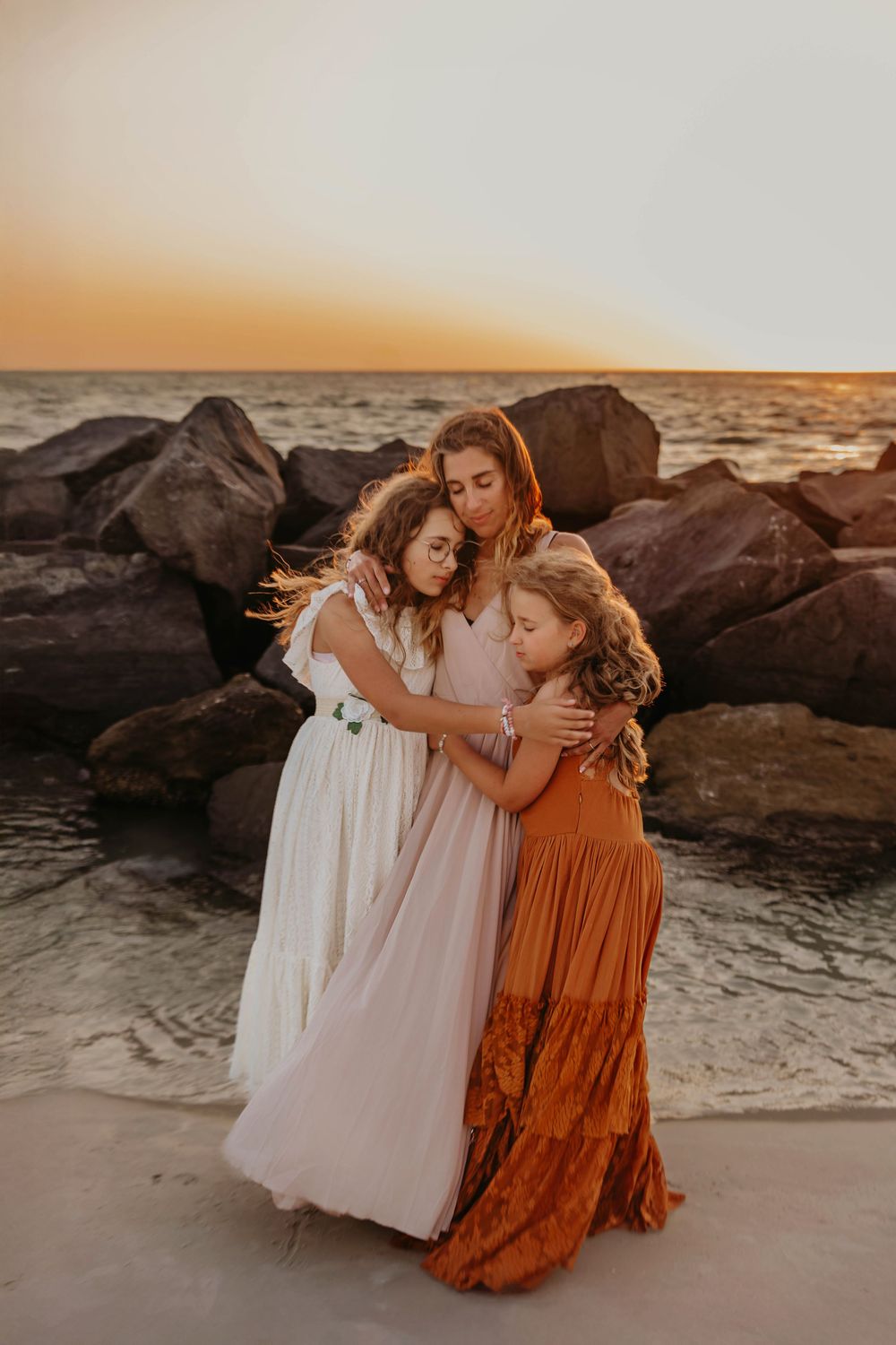 Three figures in flowing dresses embrace on a beach at sunset with dark rocks and golden light in the background.