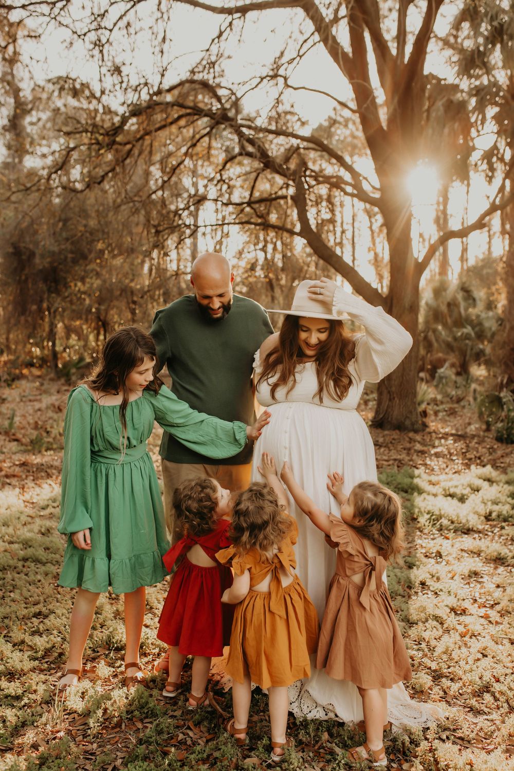 Family in coordinated outfits stands in warm sunlit forest scene during golden hour sunset.