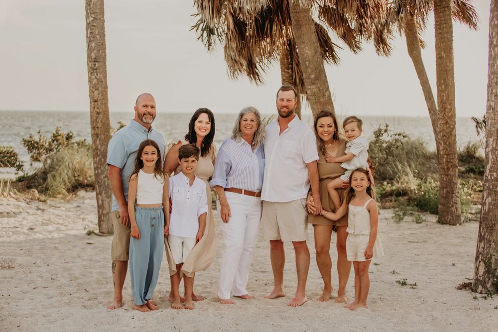 A group of eight people in coordinating beige and white outfits pose together on a beach with palm trees at sunset.