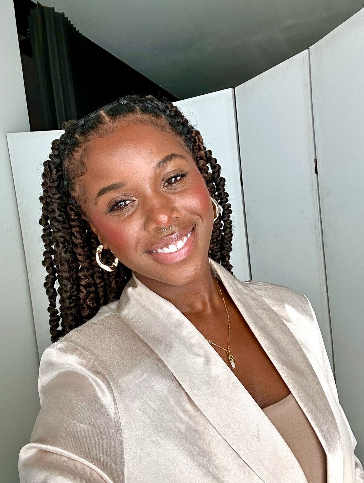 Smiling Black woman with braided hair wearing cream blazer takes selfie with hoop earrings.