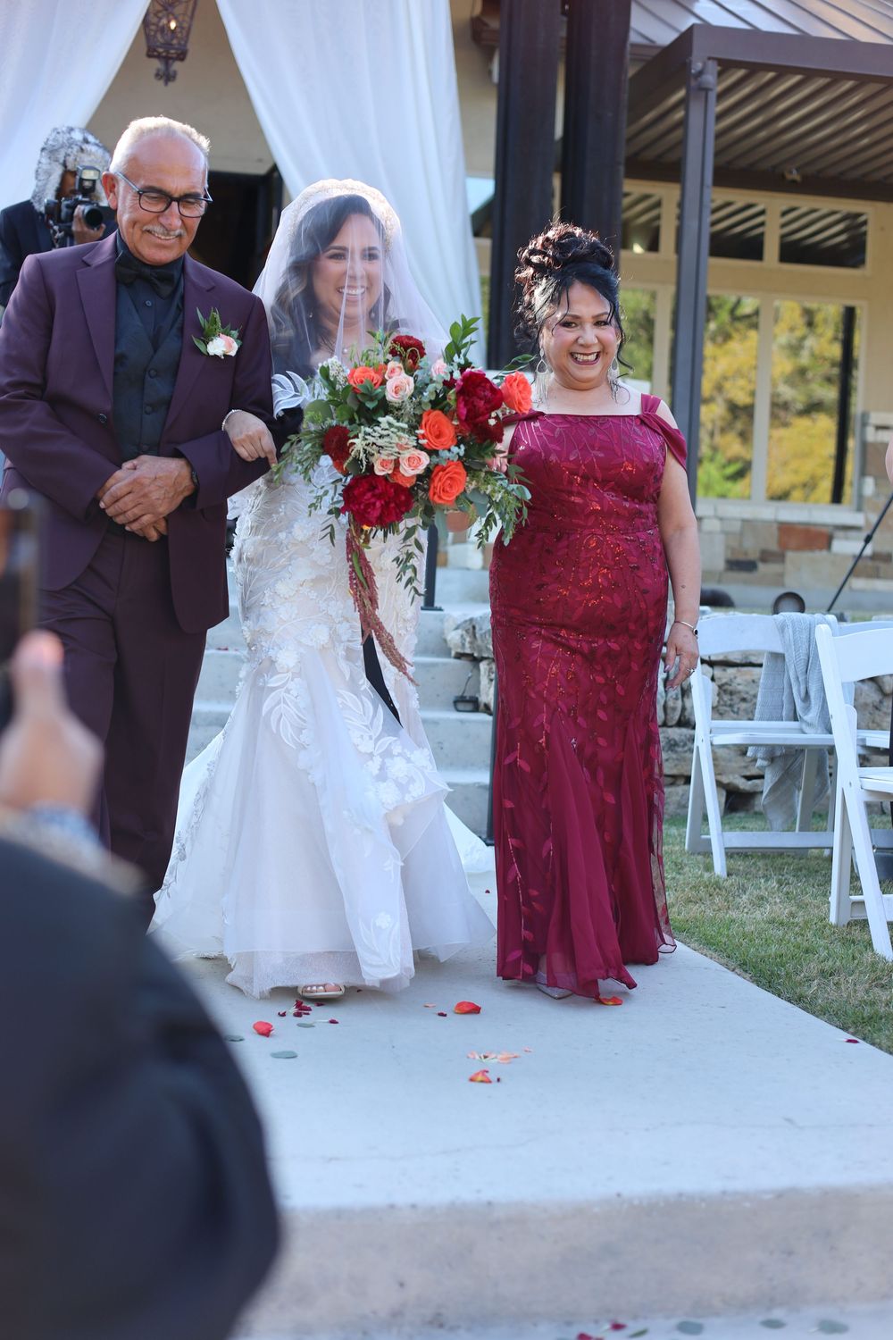 A small group walks down an outdoor aisle with red rose petals during a wedding ceremony with white chairs.