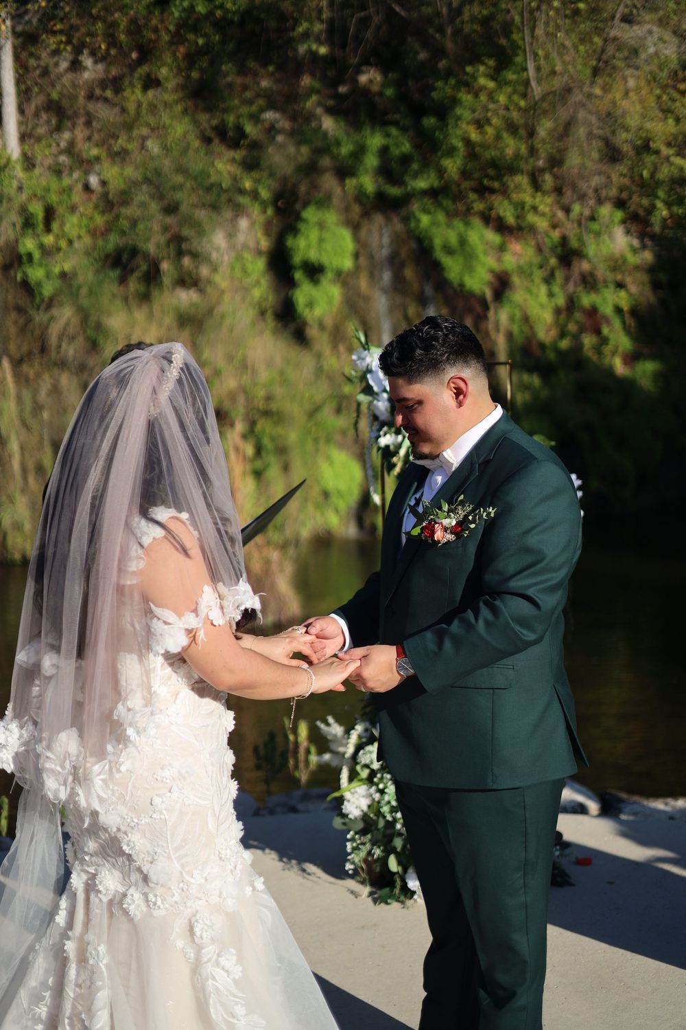 Groom in dark green suit places wedding ring on bride's finger during outdoor ceremony by a serene lake.