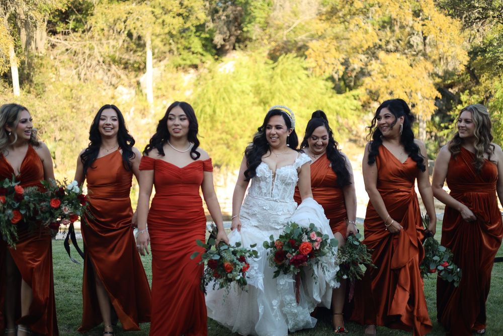 Wedding party members in burgundy dresses stand together outdoors against autumn foliage.