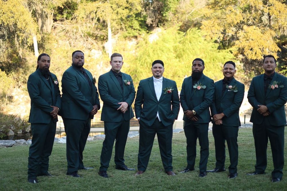 Groomsmen in dark suits pose in a line on grass with trees in the background.