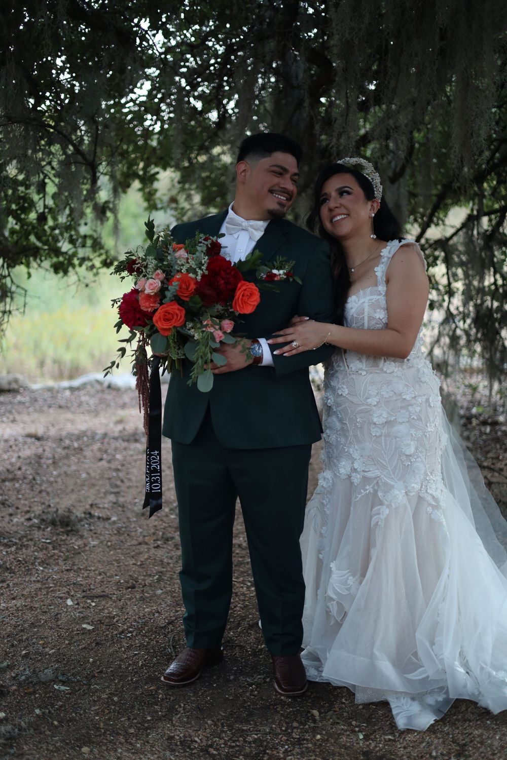 A bride and groom pose together under a tree, she in a lace gown, he in a dark suit holding a red rose bouquet.