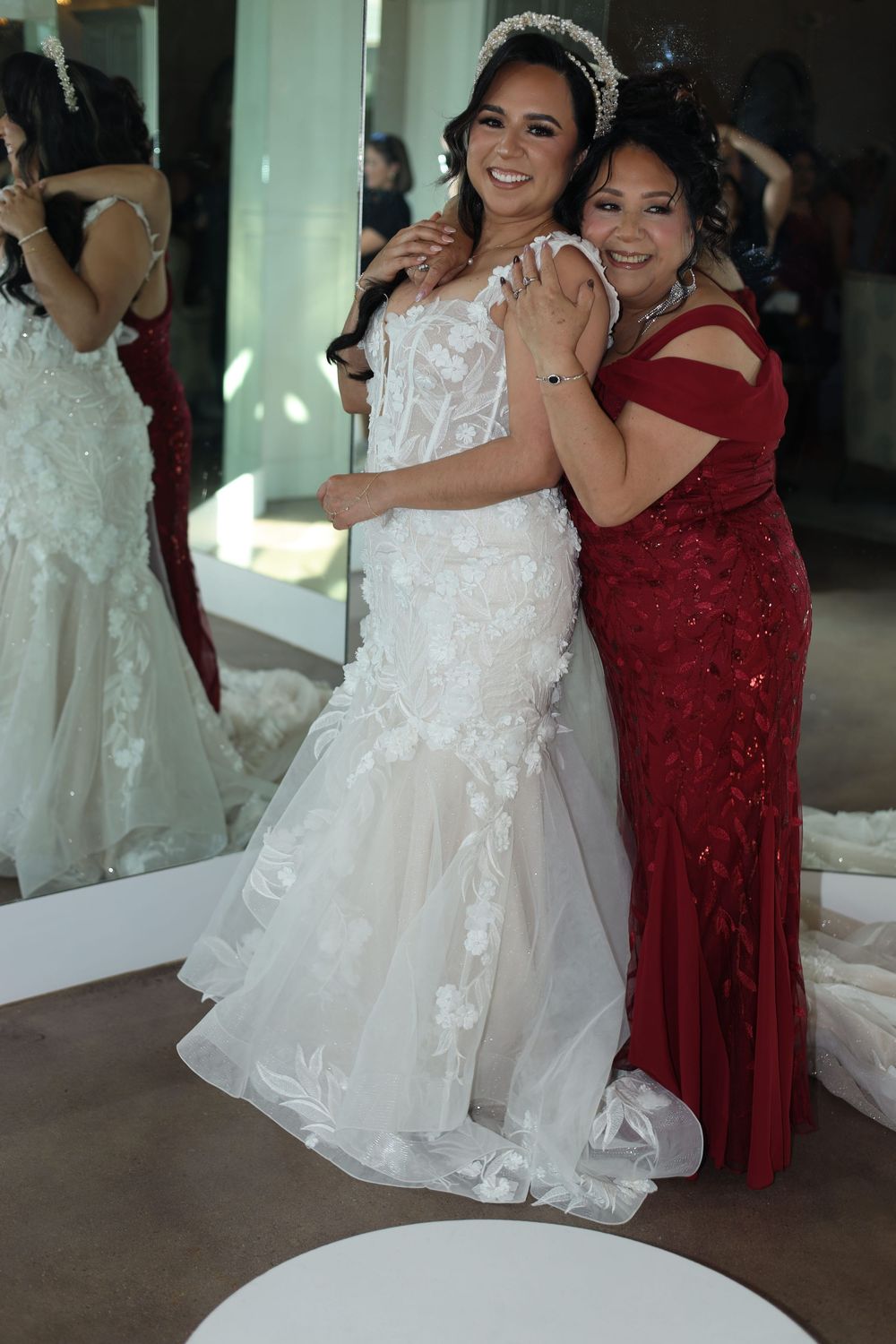 Bride in white lace wedding gown hugs woman in red dress, both smiling in bridal boutique.