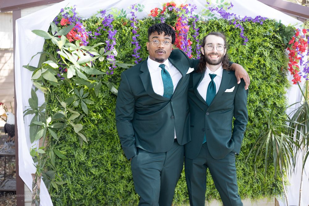 Wedding party members in dark green suits stand together in front of a lush garden wall with colorful flowers.