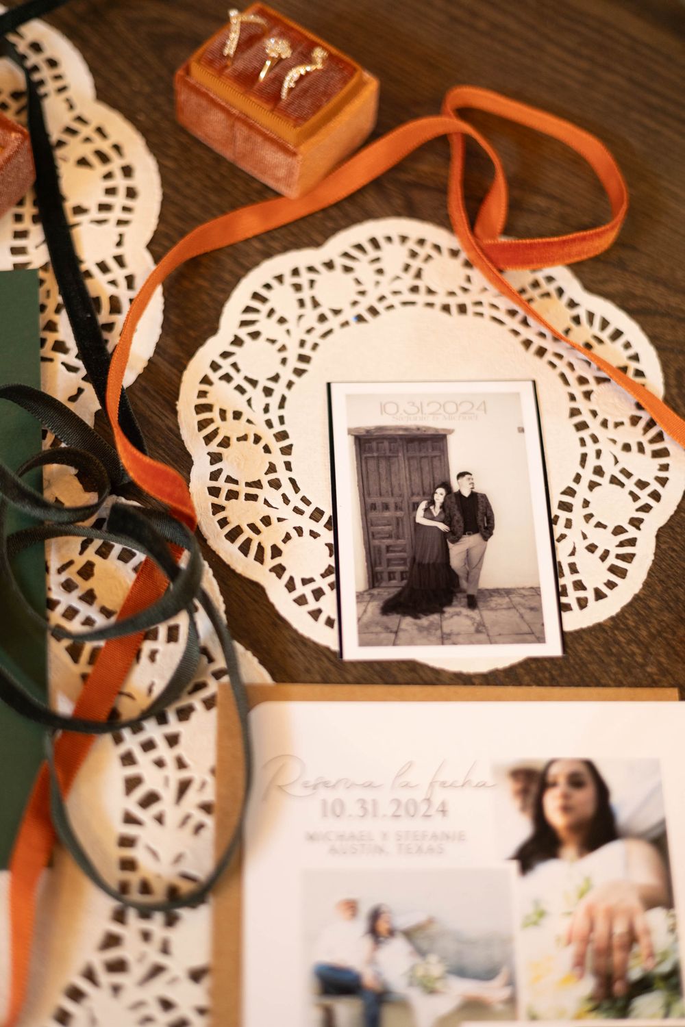 Wedding flat lay with save-the-date cards, black and white couple photo, lace doilies, orange ribbon, and ring box on wood table.