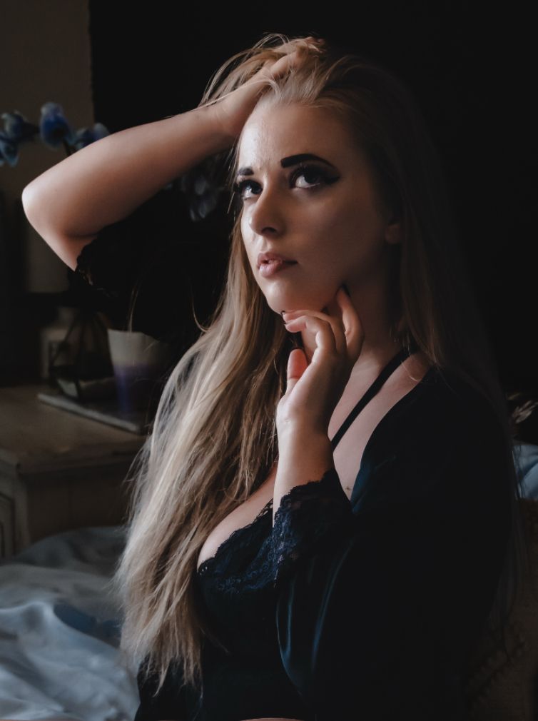 Young woman with long blonde hair in black lingerie poses thoughtfully in moody, dimly lit bedroom setting.